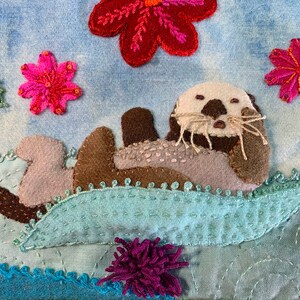 May include: A brown and gray sea otter with a white face and whiskers is floating on its back on a blue and green wave. The otter is surrounded by pink and red flowers.