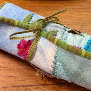 May include: A blue and green fabric roll tied with a green ribbon and decorated with a pink flower. The fabric is patchwork with different shades of blue and green.