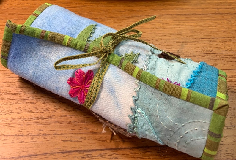 May include: A blue and green fabric roll tied with a green ribbon and decorated with a pink flower. The fabric is patchwork with different shades of blue and green.