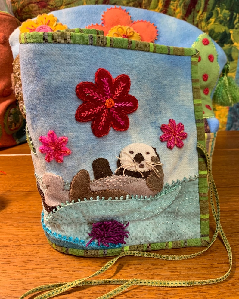 May include: A blue fabric pouch with a green and brown trim. The pouch features an embroidered sea otter, flowers, and a fuzzy purple pom-pom.