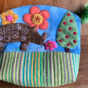 May include: A blue fabric pouch with a brown rhino, a green tree, and two pink and yellow flowers. The pouch has a striped bottom with green, blue, and yellow stripes.