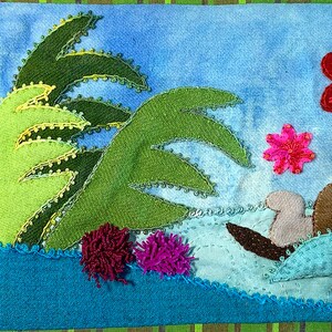 May include: A whimsical felt appliqu&eacute; depicting a sea otter swimming in turquoise water with green palm trees and pink flowers.