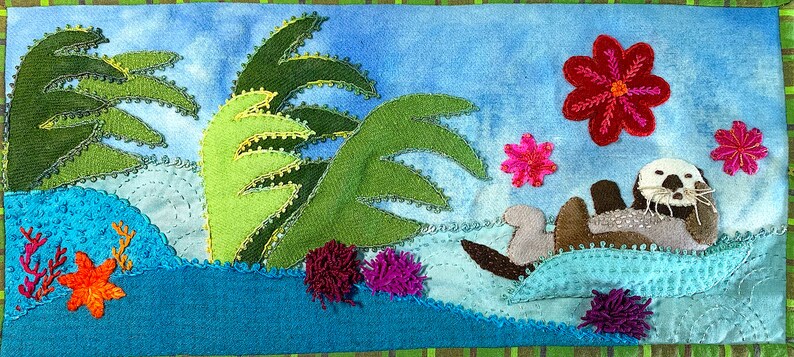 May include: A whimsical felt appliqu&eacute; depicting a sea otter swimming in turquoise water with green palm trees and pink flowers.