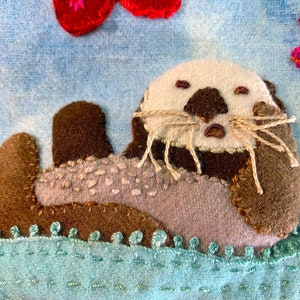 May include: A brown and gray felt sea otter with a white face and brown nose, lying on its back on a blue felt background with a green felt water border. The otter has brown felt whiskers.