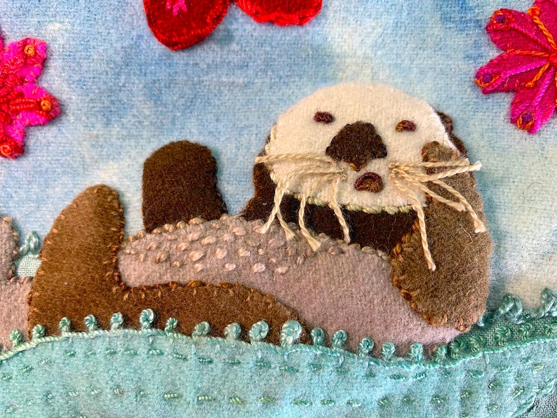 May include: A brown and gray felt sea otter with a white face and brown nose, lying on its back on a blue felt background with a green felt water border. The otter has brown felt whiskers.