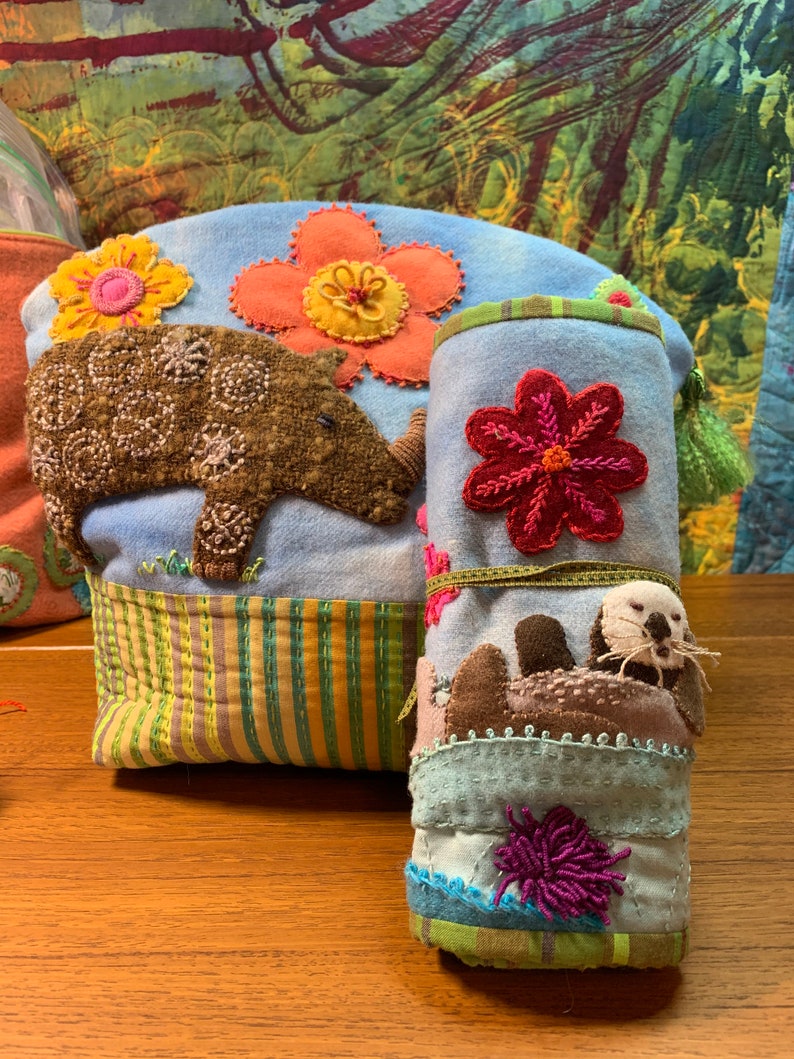 May include: Two blue fabric pouches with embroidered flowers and animals. The larger pouch has a brown rhinoceros and a yellow flower. The smaller pouch has a brown sea otter and a red flower.