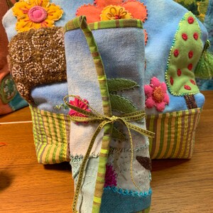 May include: A blue and green fabric roll with a brown ribbon tied around it. The fabric is decorated with colorful flowers and leaves. The roll is sitting on a wooden surface.