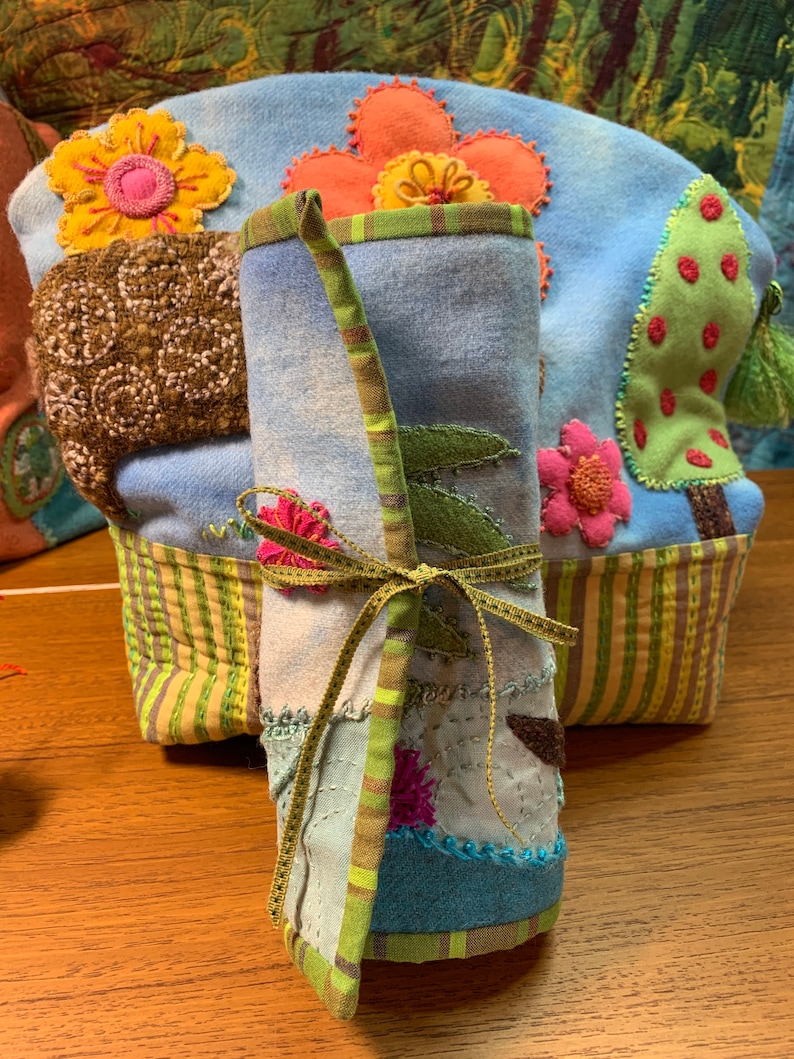 May include: A blue and green fabric roll with a brown ribbon tied around it. The fabric is decorated with colorful flowers and leaves. The roll is sitting on a wooden surface.
