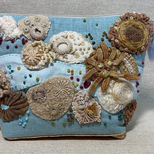 Puede incluir: Una bolsa de lino azul claro con cierre de cremallera, decorada con varios diseños bordados en tonos beige, marrón y blanco. La bolsa tiene un pequeño borla azul.