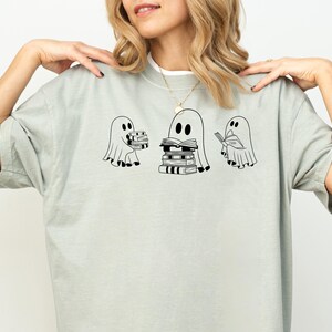 May include: A light grey t-shirt featuring a graphic of three ghosts reading books. The ghosts are outlined in black and have simple facial features. The ghost in the middle is holding a stack of books.
