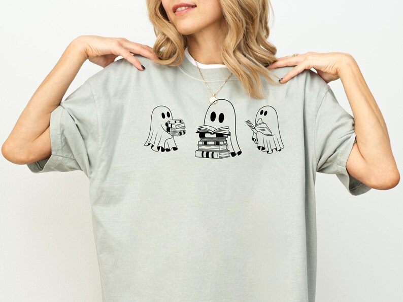 May include: A light grey t-shirt featuring a graphic of three ghosts reading books. The ghosts are outlined in black and have simple facial features. The ghost in the middle is holding a stack of books.