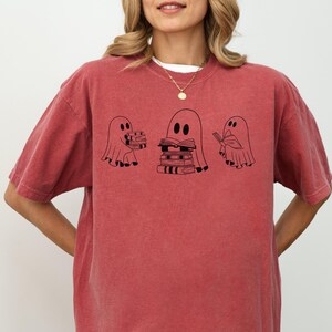 May include: A red t-shirt featuring a graphic of three ghosts holding books. The ghosts are black and white and the text on the shirt reads "Bookish Ghosts".