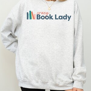 K&ouml;nnte beinhalten: Grauer Pullover mit einer bunten Grafik von gestapelten B&uuml;chern und dem Text "crazy Book Lady".