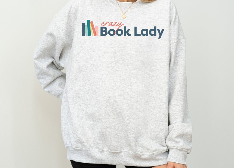 K&ouml;nnte beinhalten: Grauer Pullover mit einer bunten Grafik von gestapelten B&uuml;chern und dem Text "crazy Book Lady".