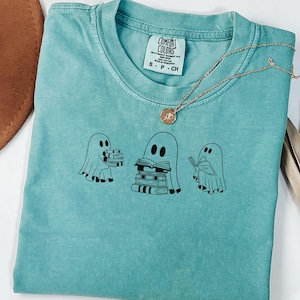 May include: A teal blue t-shirt featuring a graphic of three ghosts holding and reading books. The tag reads "Comfort Colors".