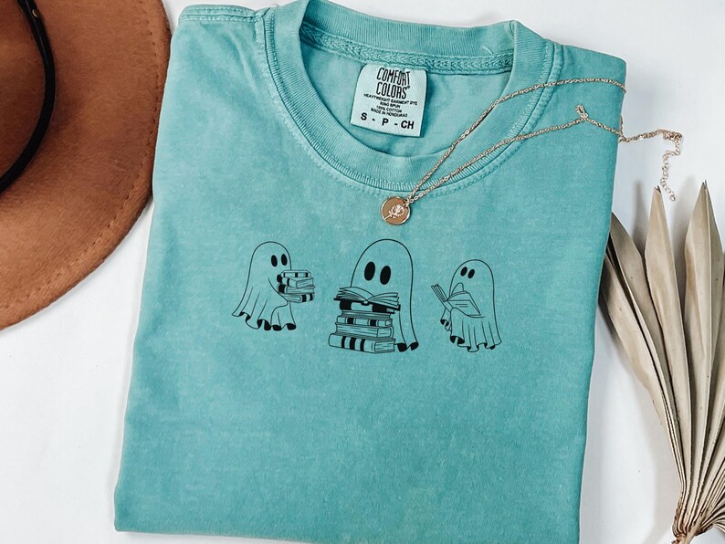 May include: A teal blue t-shirt featuring a graphic of three ghosts holding and reading books. The tag reads "Comfort Colors".