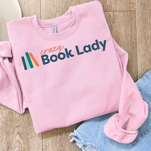 K&ouml;nnte beinhalten: Rosafarbener Pullover mit einer Grafik von bunten B&uuml;chern und dem Text "crazy Book Lady".