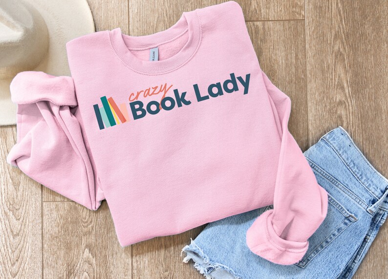 K&ouml;nnte beinhalten: Rosafarbener Pullover mit einer Grafik von bunten B&uuml;chern und dem Text "crazy Book Lady".