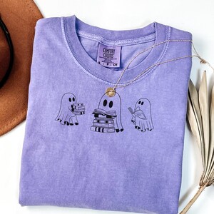 May include: A purple t-shirt featuring a design of three ghosts reading books. The label attached to the shirt reads "Comfort Colors".