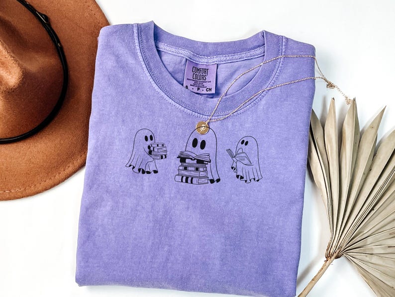 May include: A purple t-shirt featuring a design of three ghosts reading books. The label attached to the shirt reads "Comfort Colors".