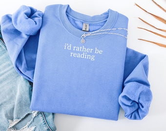 May include: A periwinkle blue sweatshirt with the phrase "I'd rather be reading" in white script. A silver necklace with a circular pendant rests on the shirt. The sweatshirt is folded with denim jeans visible.