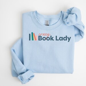K&ouml;nnte beinhalten: Ein hellblaues Sweatshirt mit dem Text "crazy Book Lady" in dunkelblau und rosa. Links neben dem Text befindet sich ein Stapel bunter B&uuml;cher.