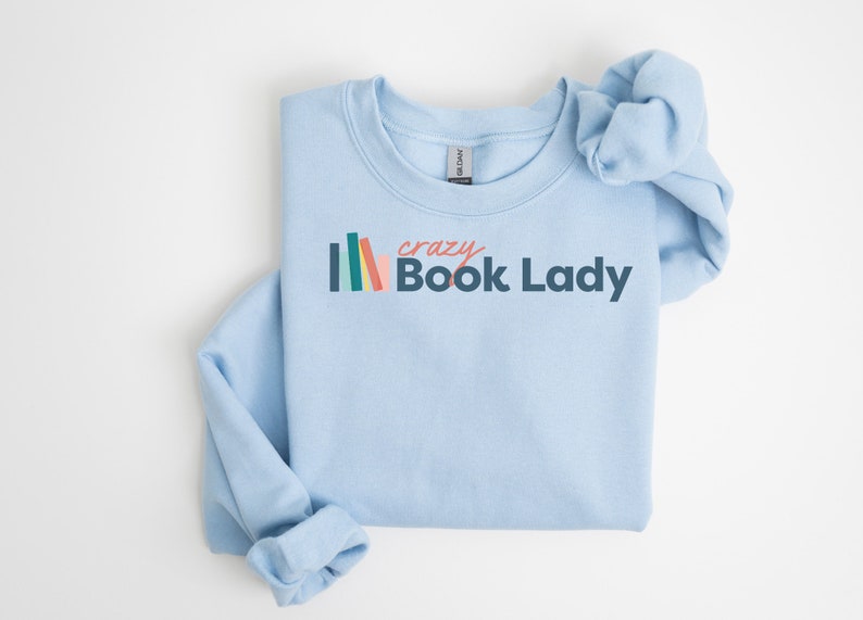 K&ouml;nnte beinhalten: Ein hellblaues Sweatshirt mit dem Text "crazy Book Lady" in dunkelblau und rosa. Links neben dem Text befindet sich ein Stapel bunter B&uuml;cher.