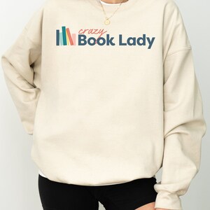 K&ouml;nnte beinhalten: Ein beiger Pullover mit einem grafischen Design aus bunten B&uuml;chern und dem Text "crazy Book Lady".