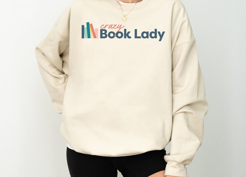 K&ouml;nnte beinhalten: Ein beiger Pullover mit einem grafischen Design aus bunten B&uuml;chern und dem Text "crazy Book Lady".