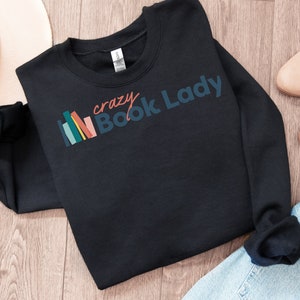 K&ouml;nnte beinhalten: Schwarzer Pullover mit dem Text "crazy Book Lady" in bunten Buchstaben und einer Grafik mit einem Stapel bunter B&uuml;cher.