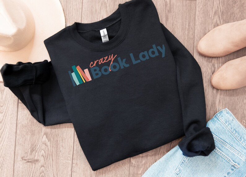 K&ouml;nnte beinhalten: Schwarzer Pullover mit dem Text "crazy Book Lady" in bunten Buchstaben und einer Grafik mit einem Stapel bunter B&uuml;cher.