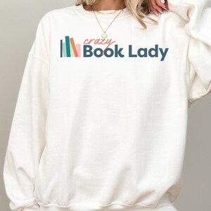K&ouml;nnte beinhalten: Wei&szlig;er Pullover mit einer bunten Grafik von gestapelten B&uuml;chern und dem Text "crazy Book Lady".