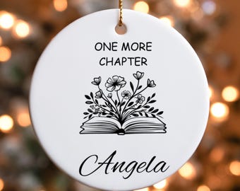 May include: White ceramic ornament with a gold string for hanging. The ornament features the text "ONE MORE CHAPTER" above a floral design over an open book. The name "Angela" is written below the book and flowers.