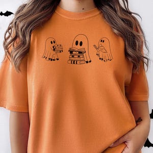 May include: Orange t-shirt featuring a graphic of three ghosts. The ghosts are holding books and one is reading a book. The text on the shirt says "Bookish Boo"
