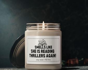 May include: A lit soy wax candle in a clear glass jar with a silver lid. The label reads "Smells Like She Is Reading Thrillers Again!" with a graphic of an open book, moon, and stars. The candle is on a dark surface.