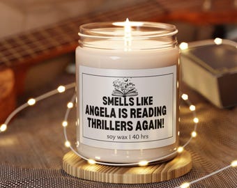 May include: A lit soy wax candle in a glass jar, resting on a wooden coaster. The label reads "Smells Like Angela Is Reading Thrillers Again!" with a book and stars graphic. The candle is surrounded by fairy lights.