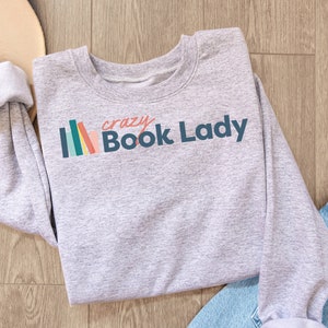 K&ouml;nnte beinhalten: Grauer Pullover mit dem Text "crazy Book Lady" in Blau und einer Grafik mit einem bunten B&uuml;cherstapel.
