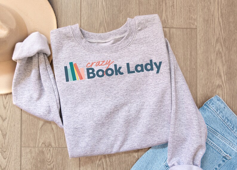 K&ouml;nnte beinhalten: Grauer Pullover mit dem Text "crazy Book Lady" in Blau und einer Grafik mit einem bunten B&uuml;cherstapel.