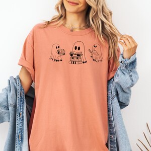May include: A peach-coloured t-shirt with a graphic of three ghosts. The ghosts are holding stacks of books. The text on the shirt says "Bookish Ghosts".