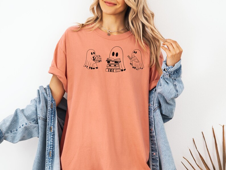May include: A peach-coloured t-shirt with a graphic of three ghosts. The ghosts are holding stacks of books. The text on the shirt says "Bookish Ghosts".