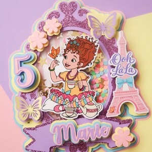 May include: A layered cake topper with a cartoon girl holding a butterfly, tea set, and Eiffel Tower. The topper is decorated with butterflies, flowers, and the name "Marie". The number "5" is also included.