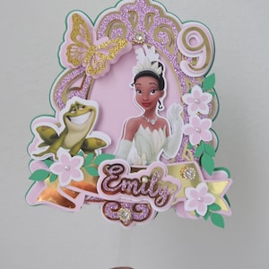 May include: A princess-themed cake topper featuring a princess illustration, a frog, and the name "Emily". The design includes pink, gold, and green accents, with a glittery finish. The number "9" is also included in the design.