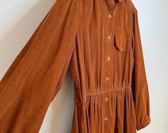 May include: A burnt orange corduroy dress with long sleeves and a button-down front. The dress features a collared neckline, a single chest pocket, and a gathered waist. The dress is hanging on a black hanger.