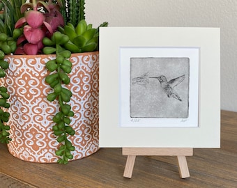 May include: A black and white etching of a hummingbird in flight, framed in a white mat and displayed on a small wooden easel. The etching is numbered 2/25 and signed by the artist.
