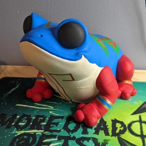 May include: A hand-painted ceramic frog figurine with blue, red, white, and gold accents. The frog has large, black eyes and a detailed design on its back.