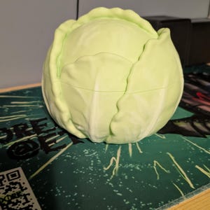 Op de afbeelding: Een lichtgroene, koolvormige container met een deksel. De container is rond en heeft een getextureerd oppervlak, dat de uitstraling van een echte kool nabootst. De container staat op een groen oppervlak.