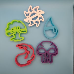 May include: Five colorful cookie cutters in the shapes of a tree, sun, water drop, flame, and skull. The cutters are in shades of green, white, blue, red, and purple. The objects are arranged on a light blue surface.