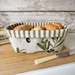 May include: A rectangular bread basket with a striped rim and olive branch pattern. The basket contains several golden-brown rolls. A butter knife rests on a wooden chopping board next to the basket.