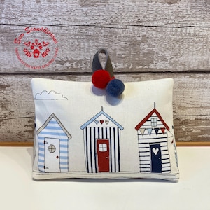 May include: White doorstop with a beach hut design. The doorstop features three blue and white striped beach huts with red roofs and red hearts. The doorstop has a white background with a blue and white trim. The doorstop has a red and blue pom-pom on top.