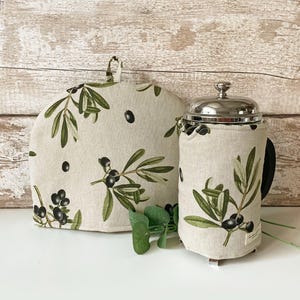 May include: A tea cosy and French press cosy set. Both are beige with a green olive branch and black olive pattern. The tea cosy has a loop handle. The French press has a silver lid and black handle.